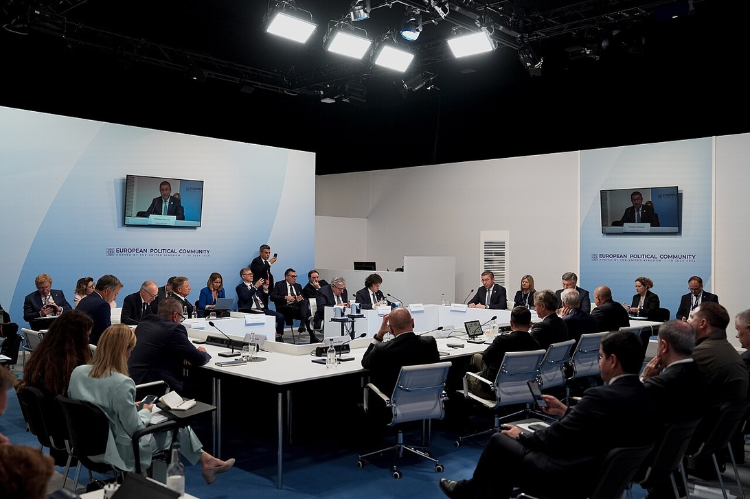 Ett mötesrum för ett politiskt toppmöte på hög nivå med böjda vita konferensbord, flera skärmar och formellt klädda politiska ledare i ett rundabordssamtal.